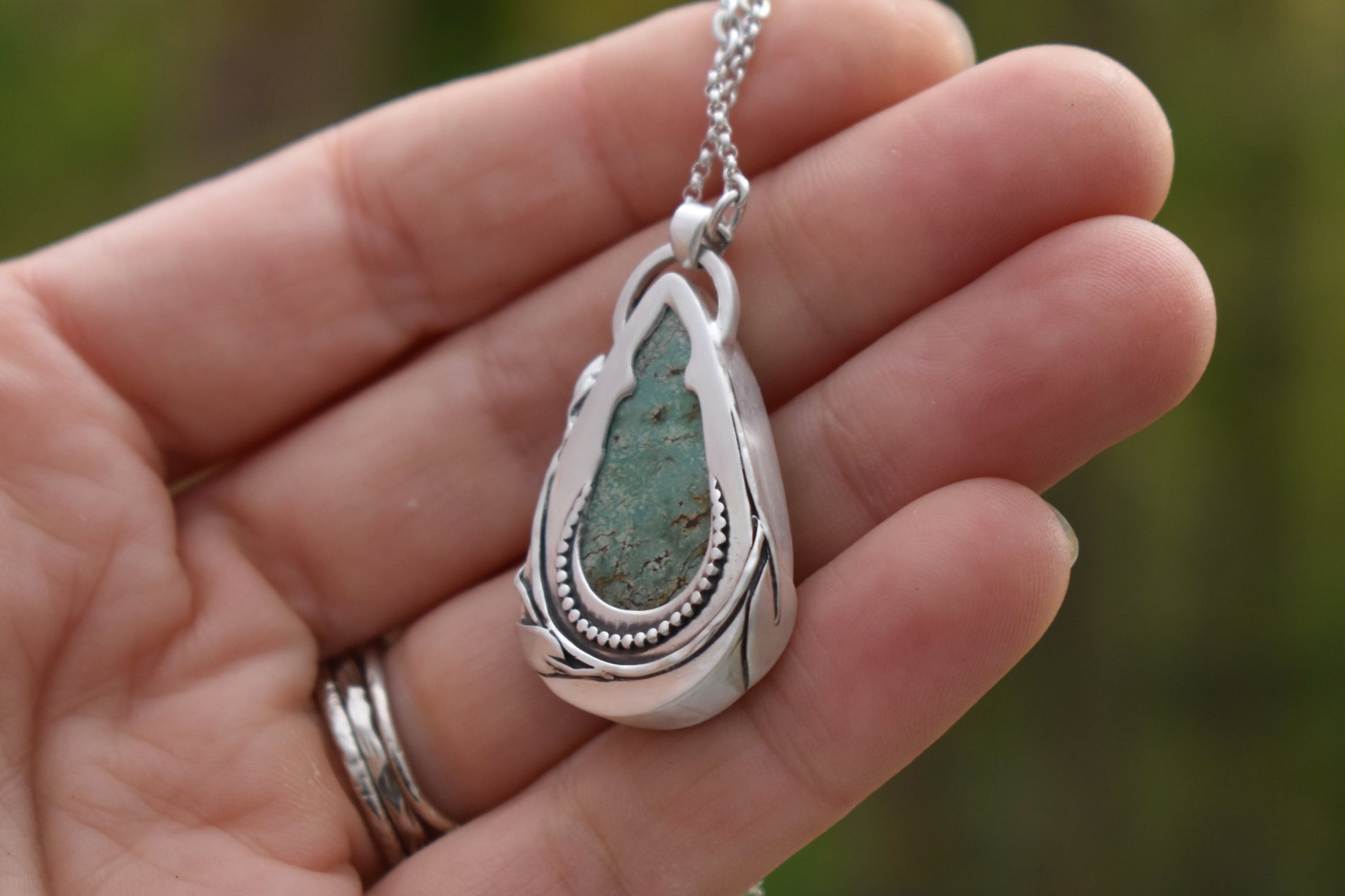 Silver pendant with green stone held in a hand against a blurred natural background