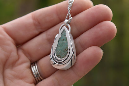 Silver pendant with green stone held in a hand against a blurred natural background